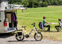 Load image into Gallery viewer, Van with a bike attached, people sitting at a table in a grassy field
