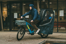 Load image into Gallery viewer, Person riding a cargo bike with a child in a covered seat on a city street.