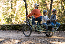 Load image into Gallery viewer, A lifestyle image showing a woman cycling a Yuba Spicy Curry+ cargo bike with two child passengers along a rural dirt track.