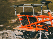 Load image into Gallery viewer, Gray metal safety bars with black padding mounted on the rear rack of an orange electric cargo bike for child protection.