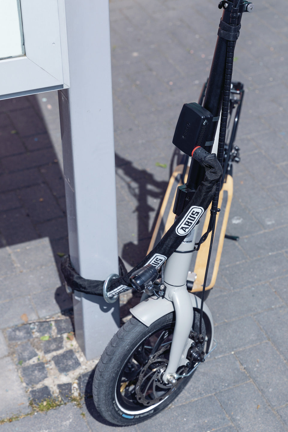 A photo of an escooter with an Abus Alarmbox 2.0 device attached. The scooter is locked to a post. 