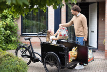 Load image into Gallery viewer, Black Bimas electric cargo trike parked outside a home, with a child sitting in the front box receiving grocery bags
