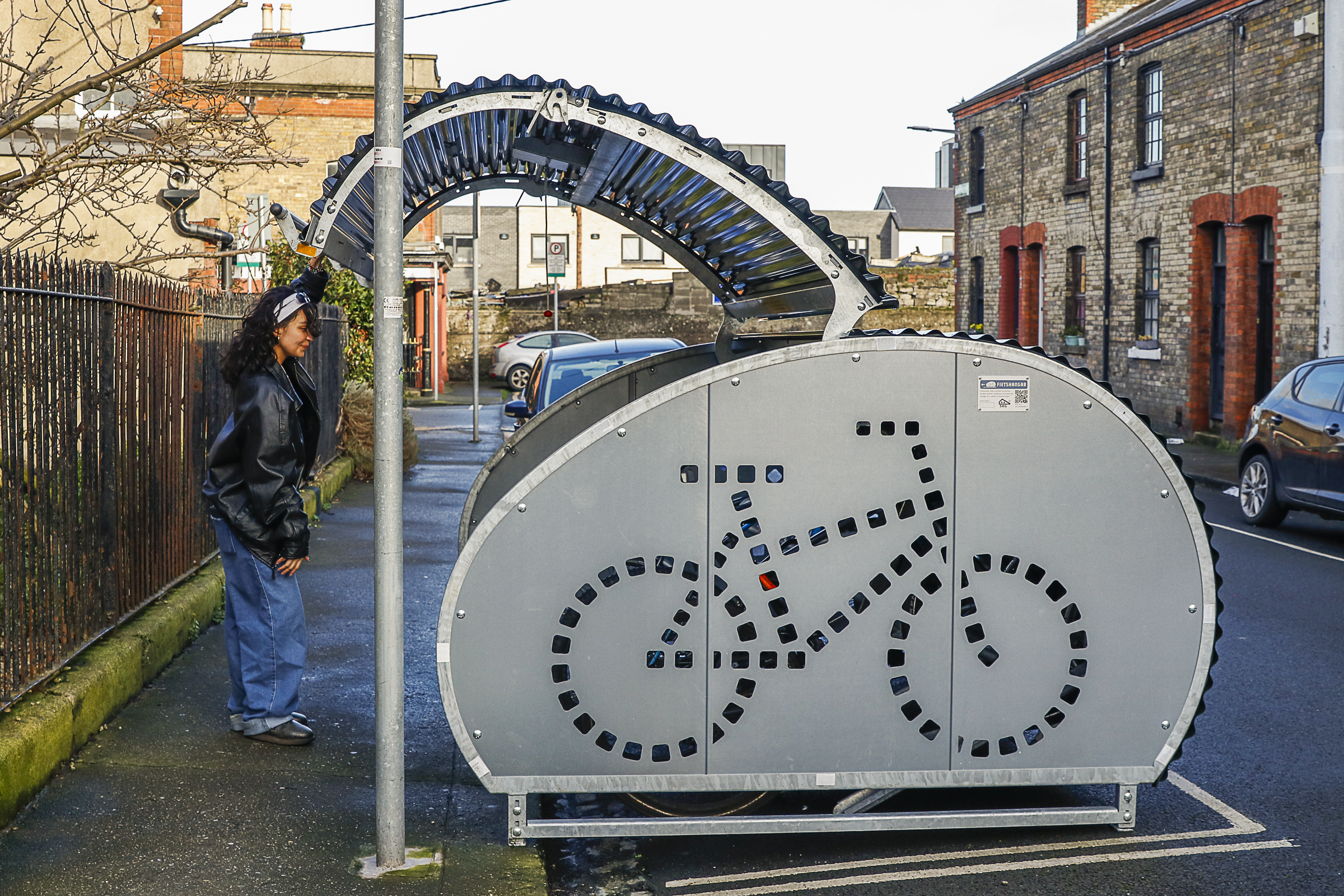 A Fietshangar bike bunker is shown on a Dublin street. A woman is opening the door of the bike bunker. 