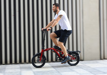 Load image into Gallery viewer, A lifestyle image showing the Dahon Curl Ei4 electric folding bike being cycled by a man in an urban setting. The frame colour is red.
