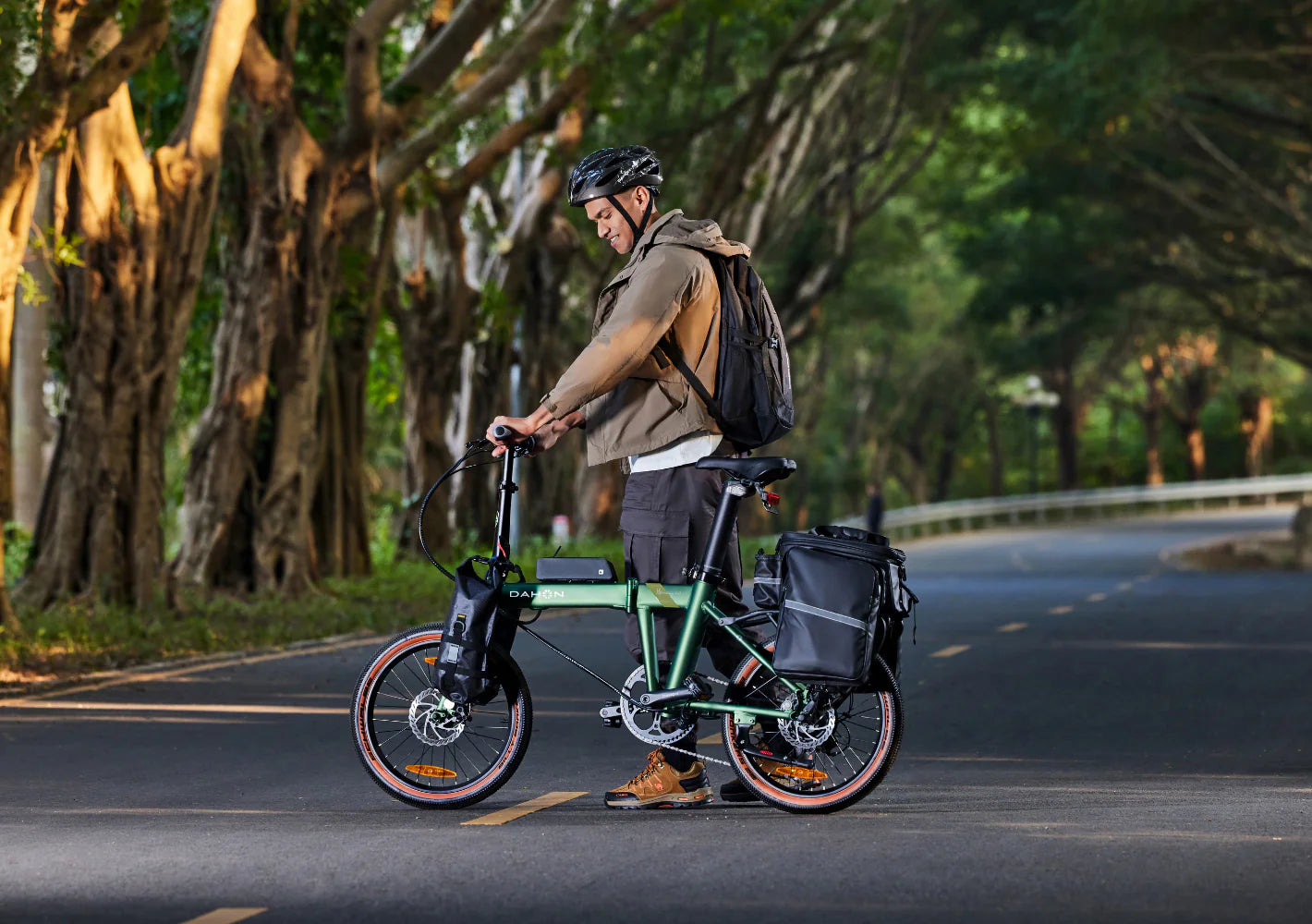 A man in a cycle helmet walking beside a green Dahon folding electric bike on a tree-lined road