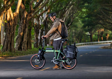 Load image into Gallery viewer, A man in a cycle helmet walking beside a green Dahon folding electric bike on a tree-lined road
