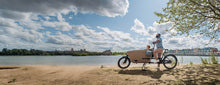 Load image into Gallery viewer, A lifestyle image of a Dolly Joy electric cargo bike parked on a beach. with a father and son standing beside the bike. 
