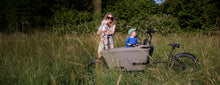 Load image into Gallery viewer, A lifestyle image of a Dolly Joy electric cargo bike parked in a grassy field, while a mother and her two children stand beside the bike. 
