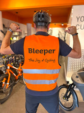 Load image into Gallery viewer, A man wearing a black bicycle helmet and a bright orange high-visibility cycling vest with 'Beeper The Joy of Cycling' printed on the back, posing in a bike shop with orange bicycles in the background.
