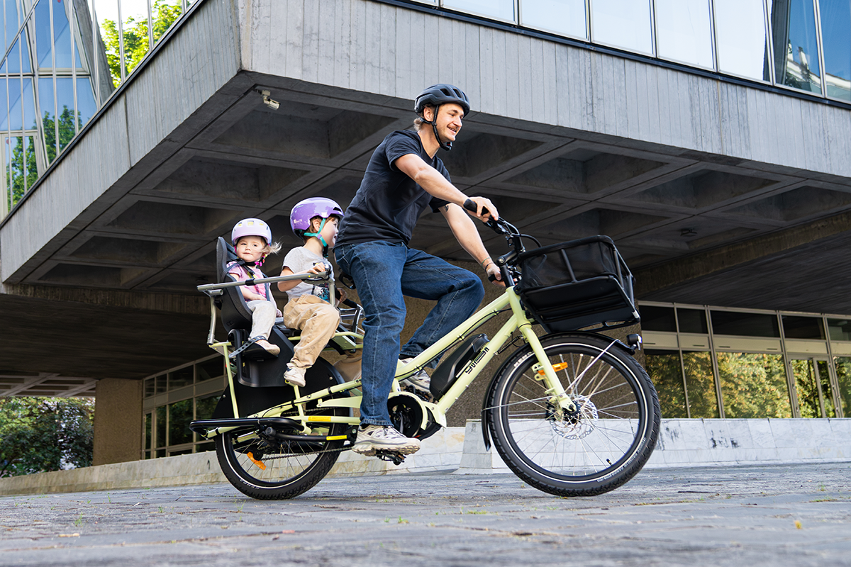 A man carries two children on the back of his Yuba Kombi EP5 electric longtail cargo bike. 