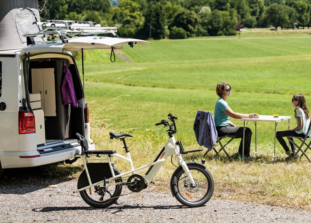 Van with a bike attached, people sitting at a table in a grassy field