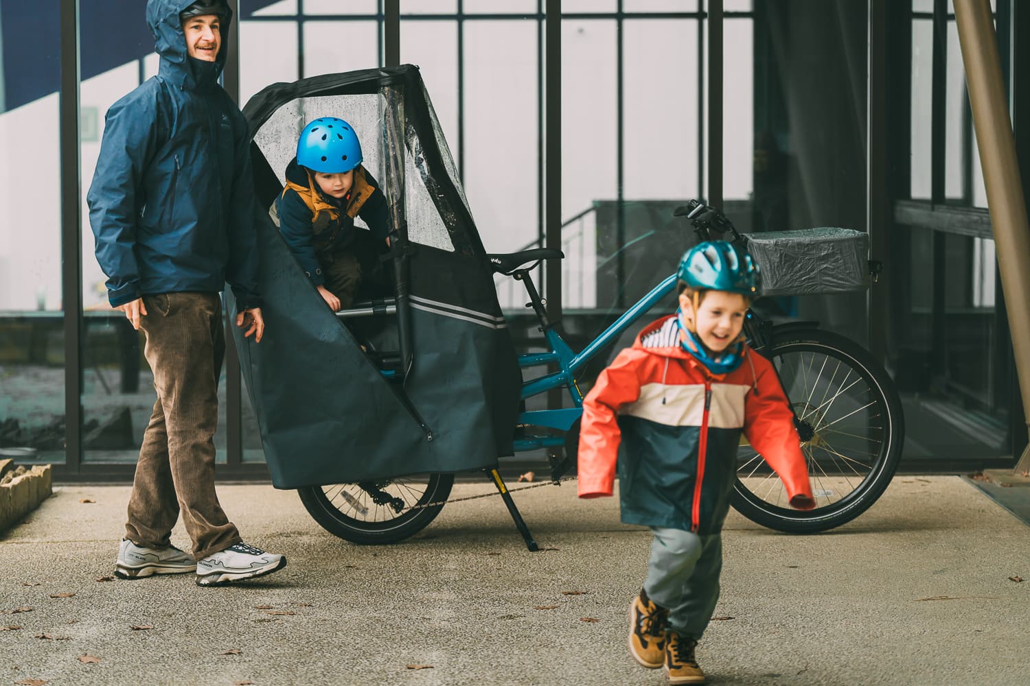 Two children with a cargo bike, one standing and one sitting in a child seat, on a sidewalk.