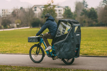 Load image into Gallery viewer, Person riding a cargo bike with a covered rear compartment on a paved path.
