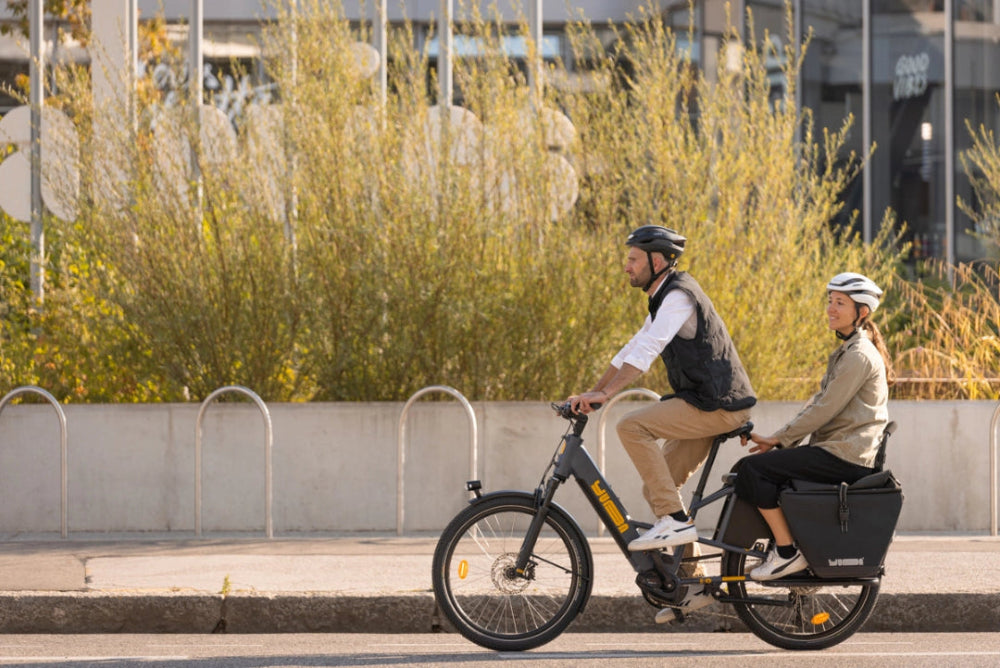 A lifestyle image of a man and a woman cycling a Yuba Boda Boda longtail electric cargo bike in an urban setting.