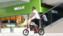 Load image into Gallery viewer, Person riding a small folding bicycle in front of a store.

