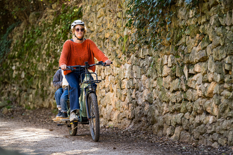 A lifestyle image showing a woman cycling a Yuba Spicy Curry+ cargo bike with two child passengers along a rural dirt track alongside an old stone wall.