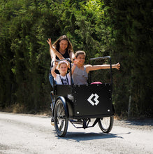 Load image into Gallery viewer, Lifestyle image of the Bimas eCargo 3.3 Premium electric cargo bike, shown in a rural setting on a gravel road with two girls as passengers in the cargo box and a woman pedalling the bike.
