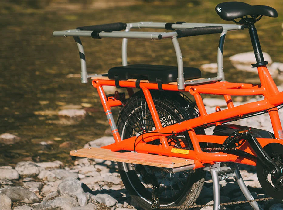 Gray metal safety bars with black padding mounted on the rear rack of an orange electric cargo bike for child protection.