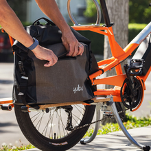 Load image into Gallery viewer, Person attaching a gray and black Yuba cargo bag to the side rack of an orange electric longtail bike for carrying gear.
